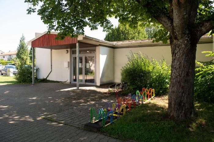 Eine kleine Kindertagesstätte mit einem überdachten Eingang, umgeben von Bäumen und Sträuchern.