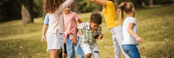 Sechs Kinder spielen zusammen in einem Park mit Gras, bilden mit ihren Armen einen Bogen.