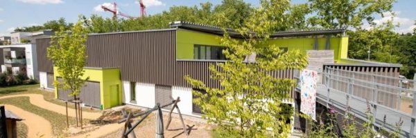 Eine moderne Kindertagesstätte mit grauen Lamellen und lindgrünen Akzenten liegt zwischen Bäumen.