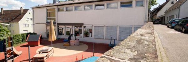 Ein Spielplatz mit Spielgeräten und Spielzeug befindet sich neben einer weißen zweistöckigen Kindertageseinrichtung.
