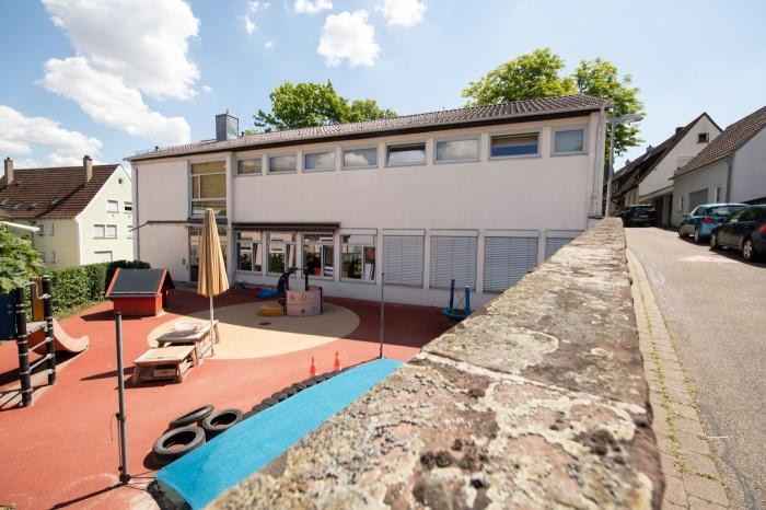 Ein Spielplatz mit Spielgeräten und Spielzeug befindet sich neben einer weißen zweistöckigen Kindertageseinrichtung.